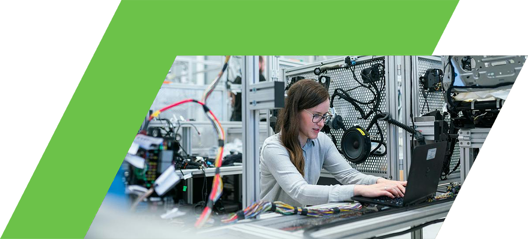 A woman works on a laptop surrounded by machinery and electrical components in a modern industrial setting.