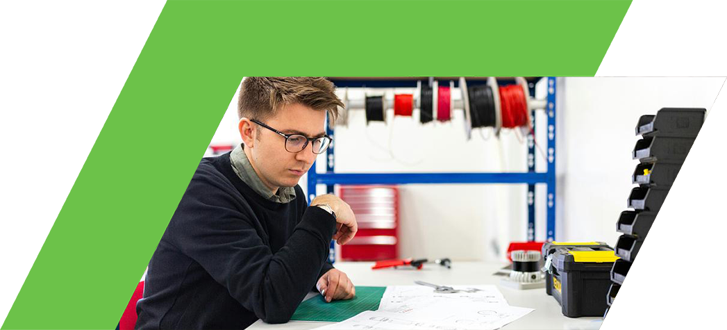 A young man is focused on a project at a workbench surrounded by tools and materials in a workshop.