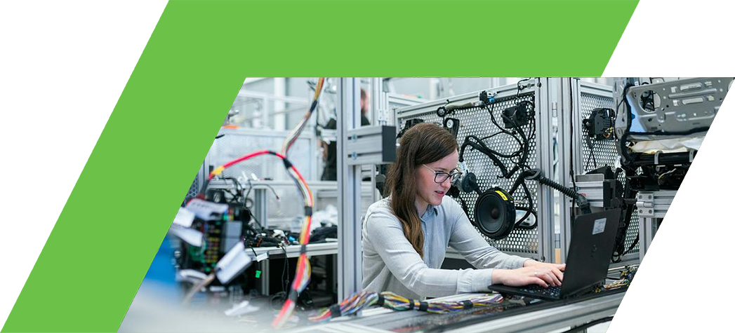 A woman works on a laptop surrounded by machinery and electronic components in a modern engineering environment.
