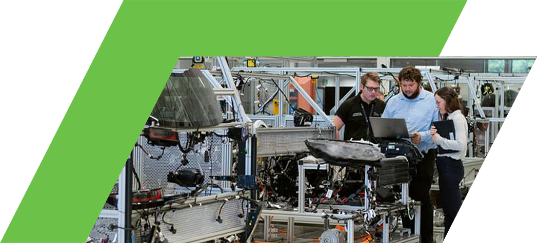 Three engineers collaborating on a project in a modern automotive workshop with various vehicle components displayed on workstations.