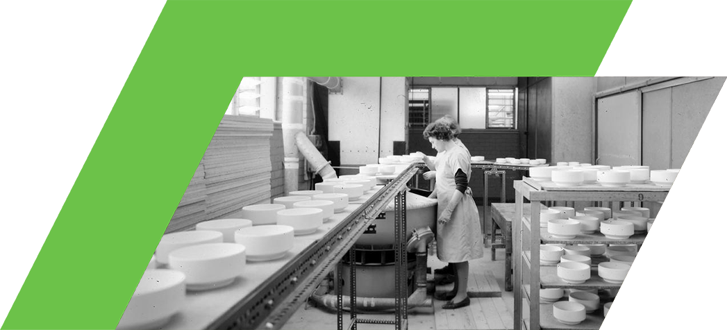 A woman in an apron works in a factory setting surrounded by rows of circular white objects on tables and shelves.