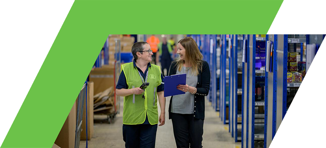 Two employees walk and talk in a warehouse aisle filled with shelves of products.