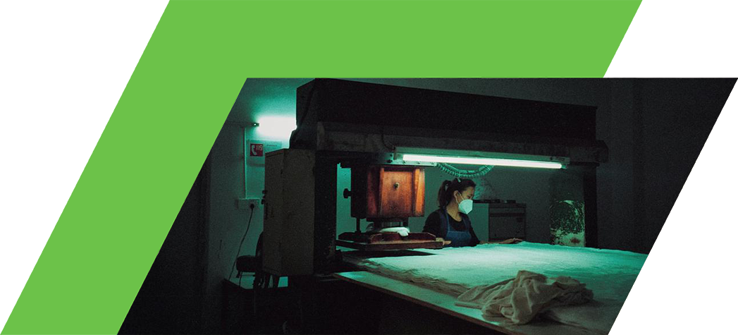 A woman wearing a mask works in a dimly lit room with a large printing press illuminated by green light.