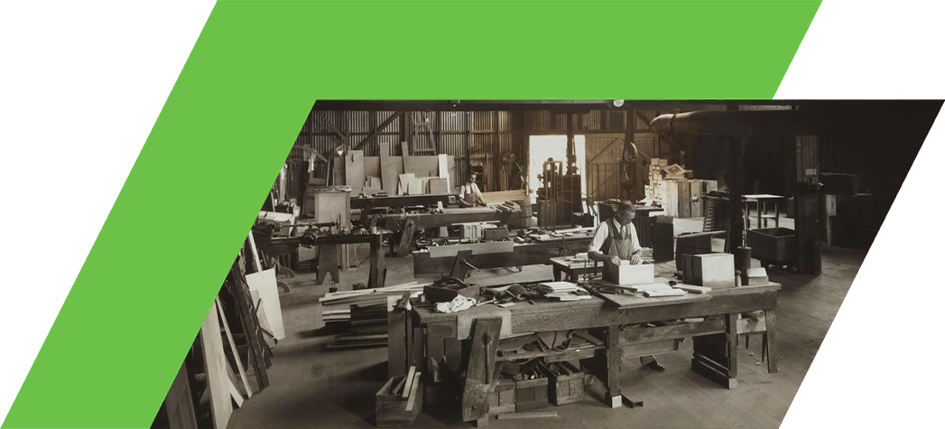 A vintage black and white photograph of a woodworking shop filled with tools, wood, and a craftsman working at a large table.