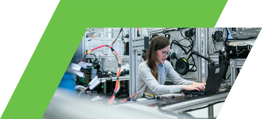 A woman works on a laptop surrounded by machinery and electronic components in a modern industrial setting.