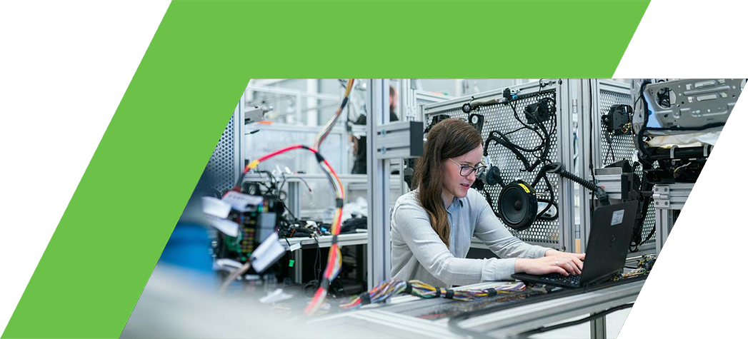 A woman works on a laptop in a high-tech manufacturing environment surrounded by machinery and electronic components.