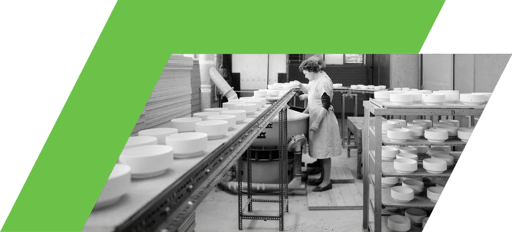 A woman in a white apron works at a table in a factory surrounded by stacked white circular objects and wooden panels.