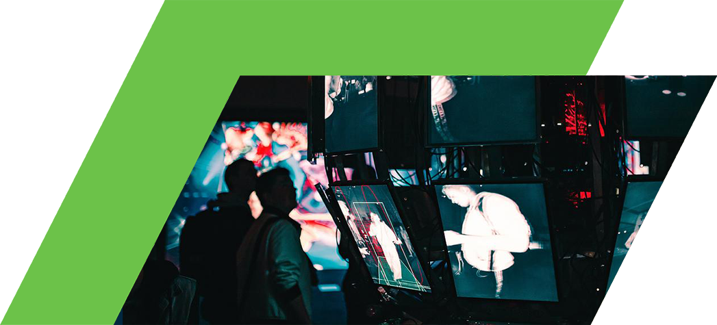 A cluster of screens displaying monochrome images in a dimly lit environment with people in the background.