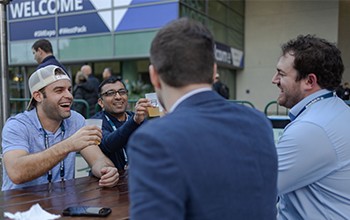 Four men sit at an outdoor table, talking and smiling, with drinks in hand. A "WELCOME" banner is visible in the background.
