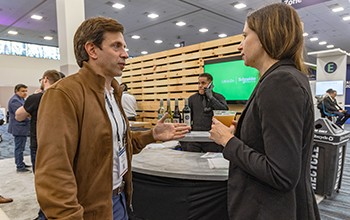  Two individuals engaged in conversation at a bustling trade show, surrounded by various booths and attendees.